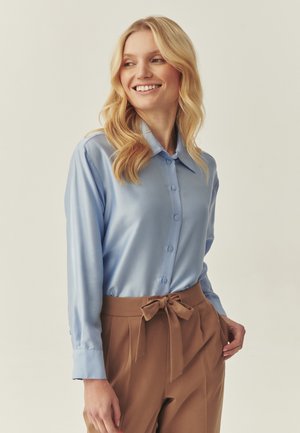 Light blue satin shirt with a pointed collar, button closure, and long sleeves, paired with brown high-waisted trousers with a tied belt.