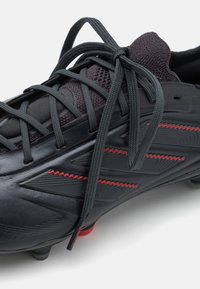 Close-up van een zwarte voetbalschoen met donkergrijze veters, rode zigzagstiksels en een gestructureerd synthetisch bovenmateriaal.