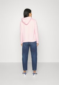 Felpa rosa chiaro con cappuccio regolabile, polsini aderenti e orlo leggermente accorciato, abbinata a jeans dritti blu medio e sneakers bianche.
