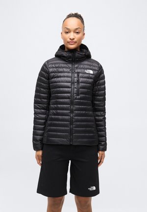 Femme portant une veste à capuche matelassée noire et un short noir, tous deux marqués du logo The North Face, debout devant un fond blanc.