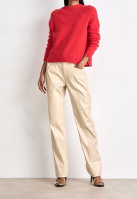 Czerwony wełniany sweter z prążkowanym dekoltem w połączeniu z beżowymi spodniami z eko skóry; modelka ma na sobie brązowe sandały na obcasie; neutralne tło.