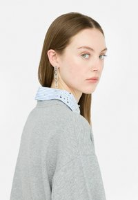 Boucles d'oreilles en argent avec un design de chaîne liée, portées avec une chemise à col rayée bleu clair sous un sweat-shirt oversized gris.