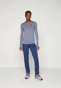 Μακρυμάνικη, ανοιχτό μπλε αθλητική μπλούζα με λογότυπο στο στήθος, συνδυασμένη με σκούρα μπλε παντελόνια και γκρι παπούτσια για πεζοπορία. Λείο υφασματικό υλικό.