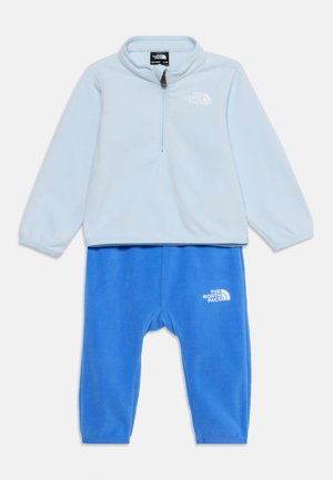 Ανοιχτό μπλε fleece μπλούζα με μακριά μανίκια και μισό φερμουάρ, και μπλε fleece παντελόνι, και τα δύο με λευκό λογότυπο The North Face στην αριστερή πλευρά.