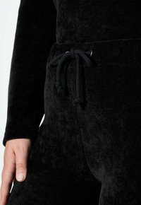 Zwarte corduroy joggers met een zachte textuur, voorzien van een trekkoord in de taille en metallic accenten. Close-up van de stof en details.