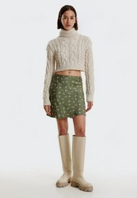 Kabelgebreide crèmekleurige cropped trui, groene minirok met bloemenpatroon en beige kniehoge laarzen met dikke zolen. Eenvoudig en modern ontwerp.