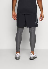 Shorts de sport noirs sur des leggings de compression gris, associés à des baskets blanches et grises. Les shorts ont un accent réfléchissant sur le côté.