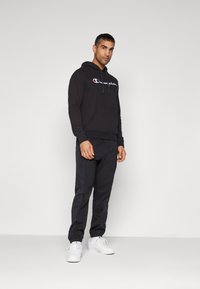 Hoodie noir avec logo, doté de cordons et d'une poche kangourou. Associé à un pantalon de survêtement noir, le tout en tissu doux. Des baskets blanches complètent le look.