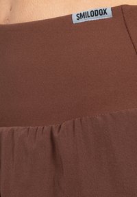 Bruna träningsshorts med en slät, texturerad tyg, med en rynkad nederkant och en etikett med texten "SMILODOX" vid midjan.