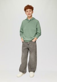 Groene hoodie met een horizontale naad, gecombineerd met losse grijze jeans en witte sneakers, staand tegen een effen achtergrond.