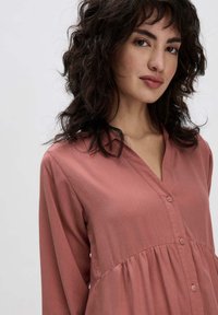Blusa color ruggine a maniche lunghe con scollatura a V, chiusura frontale con bottoni e vita arricciata, realizzata in un tessuto liscio e leggero.
