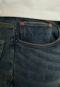 Jeans en denim bleu foncé avec une finition déchirée. Présentent des rivets en cuivre et une poche avant. Tissu en coton avec un tissage texturé.