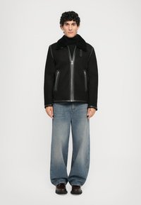 Svart jacka med fårskinnskrage, dragkedjestängning och två frontfickor, i kombination med lösa blå jeans och mörka skor.