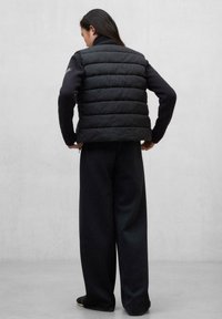 Chaleco negro de plumas con diseño acolchado horizontal, llevado sobre un suéter oscuro. El chaleco cuenta con un cuello alto y se combina con pantalones negros holgados.