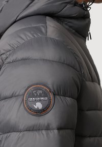 Grey padded jacket with a smooth texture, featuring a round black and orange logo patch on the left shoulder and horizontal quilting pattern.