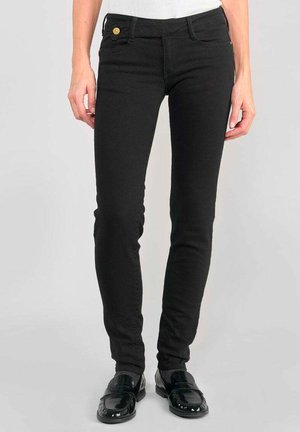 Jean noir ajusté avec cinq poches, fermeture à bouton et un tissu lisse et extensible. Porté avec des mocassins noirs brillants.
