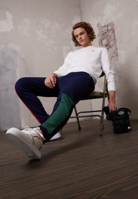 Tumesinised spordipüksid, millel on rohelised detailid ja punased külgribad, on selga pandud valgete tossudega. Mudel istub toolil, hoides käes väikest musta kotti.