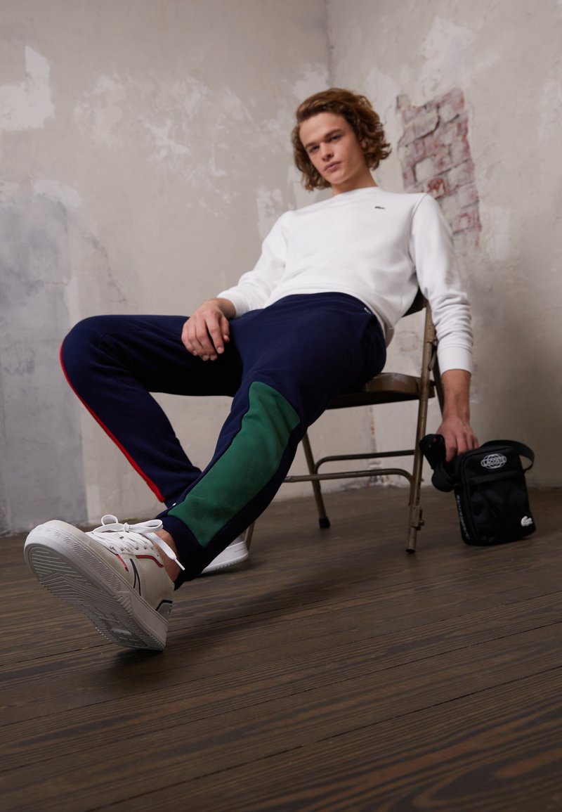 Pantalons de survêtement navy avec des accents verts et des bandes latérales rouges, portés avec des baskets blanches. Le modèle est assis sur une chaise, tenant un petit sac noir.