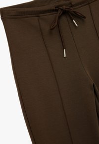 Los pantalones de chándal marrones tienen una cintura con cordón, una textura suave y detalles cosidos a lo largo del costado. Las puntas de metal en el cordón realzan el diseño.