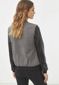 Chaqueta bomber con un cuerpo de punto gris y mangas de cuero negro, con dobladillo y puños acanalados. Diseño sencillo sin logotipos ni adornos visibles.