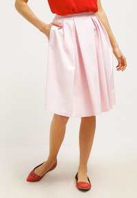 Gonna plissettata rosa in cotone, lunghezza al ginocchio, con tasche laterali e tessuto liscio. Abbinata a scarpe basse rosse.