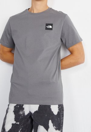 Osoba nosząca gładką szarą koszulkę z krótkim rękawem z małym czarno-białym logo The North Face na klatce piersiowej oraz czarno-białe spodnie w wzory.