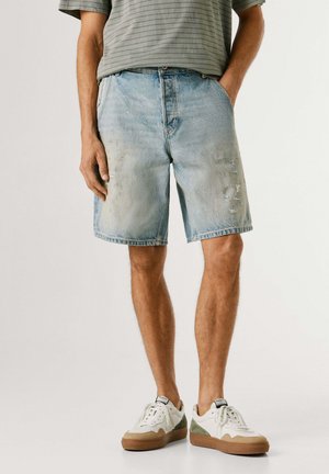 Mann trägt hellblaue zerrissene Jeansshorts, gestreiftes graues T-Shirt und weiße Turnschuhe mit braunen Sohlen und steht vor einem schlichten Hintergrund.