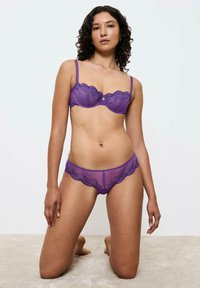 Fioletowy koronkowy biustonosz z falbaniastymi krawędziami, z cienkimi ramiączkami i detalem w kształcie otworu. Pasujące przezroczyste fioletowe majtki z podobnym falbaniastym wzorem.