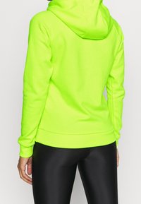 Sudadera verde neón con capucha ajustable, puños y dobladillo elásticos, con una textura suave y una silueta ajustada, combinada con leggings negros.