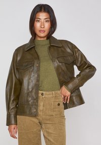 Giacca di pelle marrone con design collerato, bottoni a pressione e due tasche sul petto, indossata sopra una camicia verde oliva e pantaloni di corduroy color sabbia.