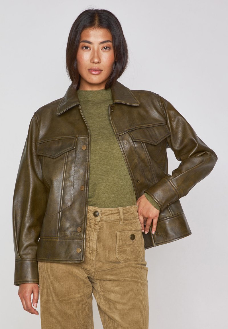 Giacca di pelle marrone con design collerato, bottoni a pressione e due tasche sul petto, indossata sopra una camicia verde oliva e pantaloni di corduroy color sabbia.