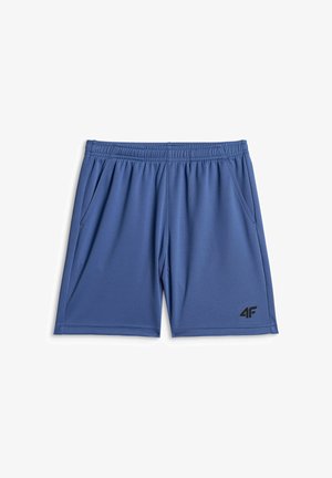 Jeune garçon debout et souriant, portant un t-shirt gris à motifs, un short bleu, des chaussettes blanches à rayures noires et des baskets noir et blanc.