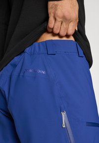 Plave vanjske hlače s glatkom teksturom, s diskretnim džepom na patentni zatvarač i logotipom marke. Struk sadrži petlje za remen.