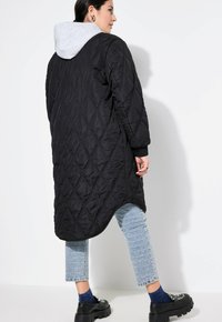 Cappotto trapuntato nero con cappuccio grigio, design lungo e polsini a coste. Indossato sopra pantaloni blu con motivo e scarpe nere ingombranti.