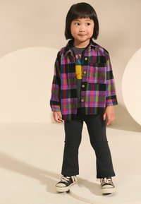 Chemise en flanelle à carreaux multicolores avec des motifs noirs, violets, verts et roses ; t-shirt graphique gris ; leggings noirs ; baskets noires avec semelles blanches.