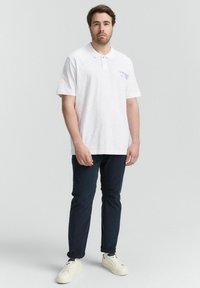 Witte poloshirt van katoen, met korte mouwen en een knoopsluiting met drie knopen. Heeft een lichtblauw logo op de linkerkant van de borst. Gecombineerd met marineblauwe broek.