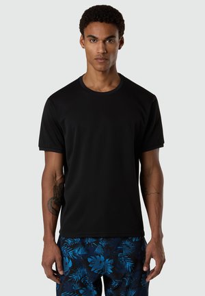 Uomo con capelli corti e ricci che indossa una maglietta nera semplice e pantaloncini blu con un motivo scuro a foglie tropicali, in piedi davanti a uno sfondo uniforme.