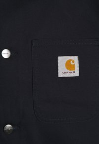 Dunkelblauer Canvas-Stoff mit einer Fronttasche. Verfügt über einen silbernen Druckknopf und ein weißes Etikett mit einem gelben Logo und dem Text "Carhartt".