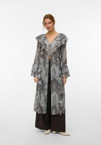 Femme portant un long cardigan transparent à motifs avec des volants, sur un pantalon large foncé et des chaussures de couleur claire, debout devant un fond uni.