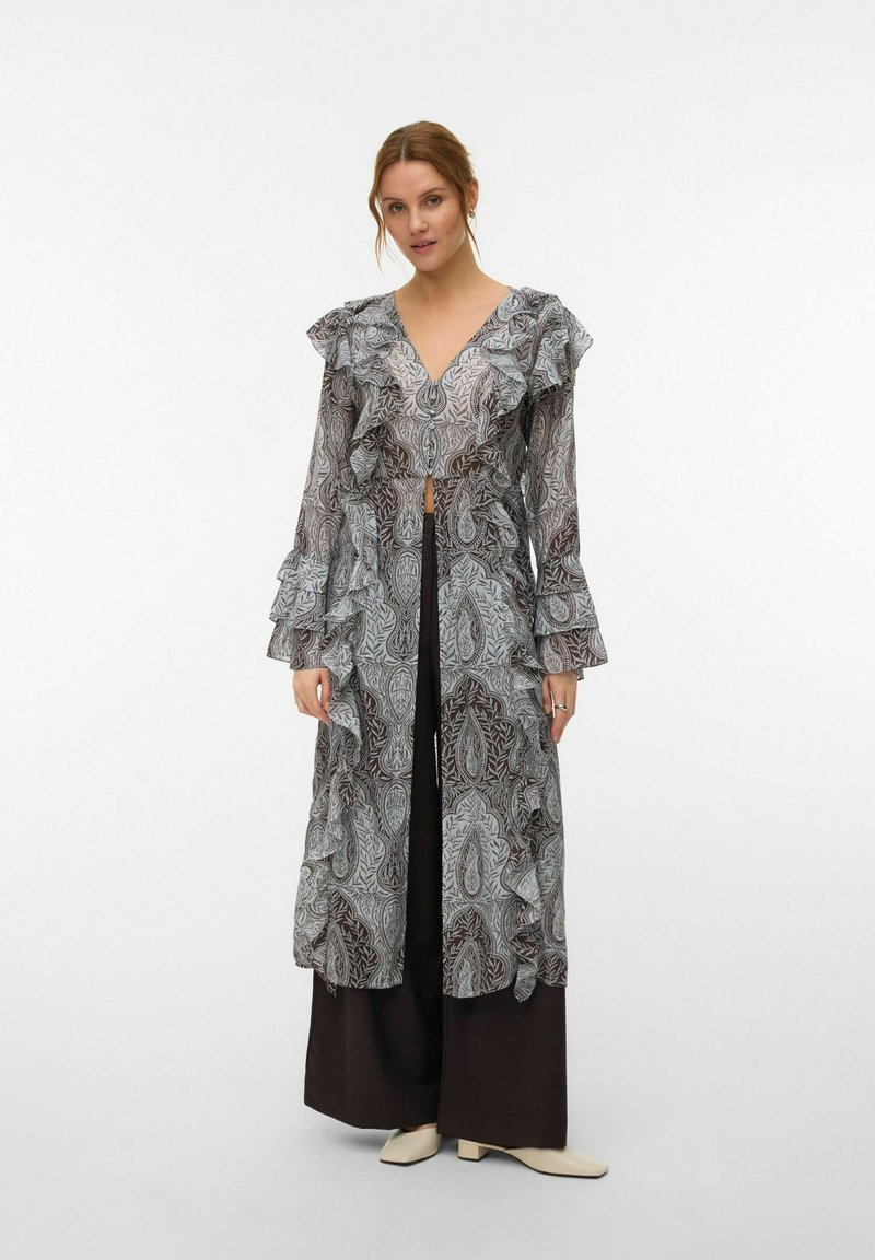 Femme portant un long cardigan transparent à motifs avec des volants, sur un pantalon large foncé et des chaussures de couleur claire, debout devant un fond uni.