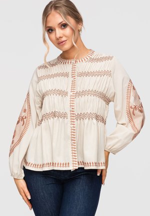 Femme portant un chemisier crème avec une broderie marron sur les manches et le devant, associé à un jean bleu foncé, debout devant un fond uni.