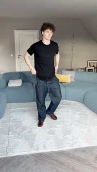Musta mynkkipaita, löysä tummansinisistä farkuista, joissa on nauha, ja ruskeat kengät, seisoo pehmeällä valkoisella matolla vaaleassa huoneessa.