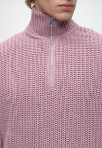 Personne portant un pull tricoté violet clair avec un col montant côtelé et une fermeture éclair argentée à l'avant, partiellement fermée.