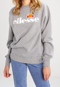 Grå sweatshirt med rundad nacke och långa ärmar. Har en vit "ellesse"-logga och orange accent. Bärs med blå jeans.