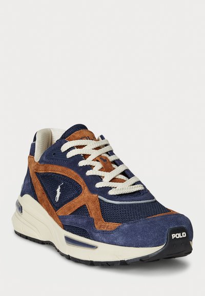 Zapatilla de deporte azul marino y marrón con materiales de ante y malla, cordones blancos y suela gruesa, que presenta un logotipo en el lateral y el talón.