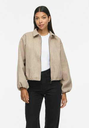 Femme aux longs cheveux foncés portant une veste beige zippée sur un haut blanc et un pantalon noir, debout devant un fond blanc uni.