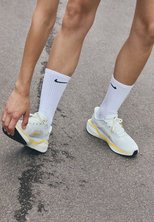 Jambes portant des chaussures de course blanches et jaunes et des chaussettes Nike blanches sur l'asphalte, avec une main touchant le talon de la chaussure gauche.