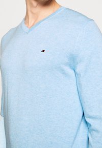 Helles blaues V-Neck-Pullover aus weichem Stoff, mit einem kleinen Logo in rot, weiß und blau auf der linken Brust.