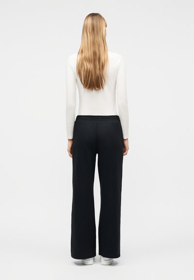 Γυναίκα με μακριά ξανθά μαλλιά, φορώντας λευκό μπλουζάκι με μακριά μανίκια, μαύρο παντελόνι φαρδύ, και λευκά παπούτσια, στέκεται με την πλάτη προς τα εμπρός σε μία απλή φόντο.