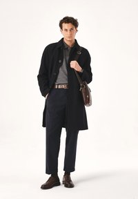 Abrigo de lana negro sobre un polo gris, pantalones navy y zapatos de cuero marrón. Incluye un bolso de hombro marrón. Diseño simple y estructurado.